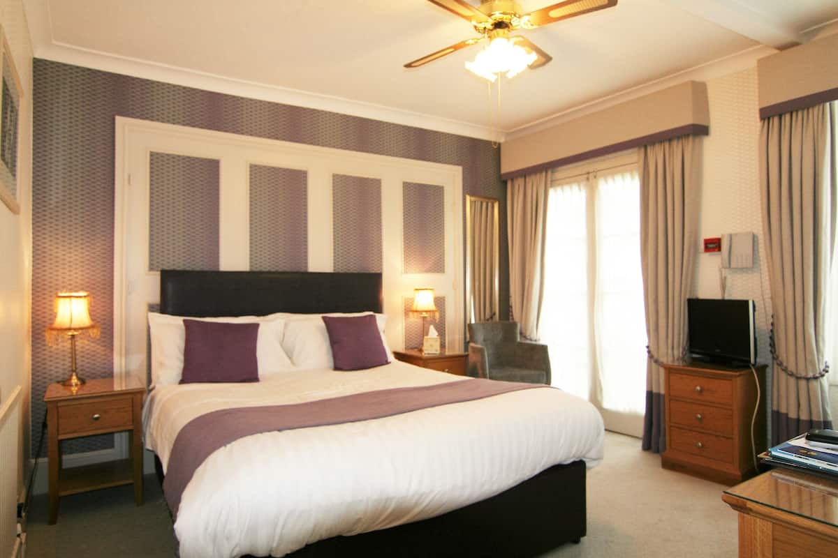 A hotel room with a large bed, a ceiling fan, a television, a desk, and a chair.