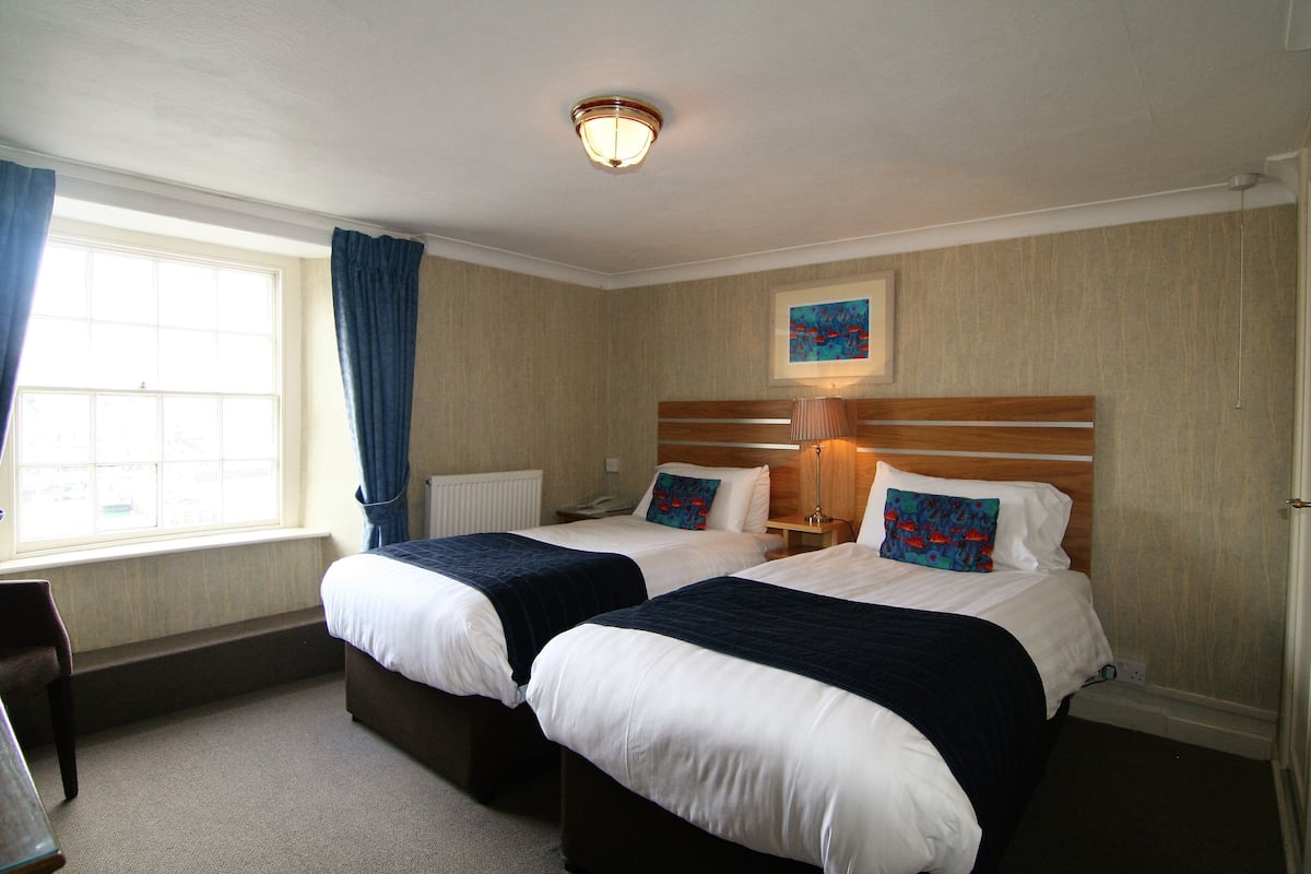 A hotel room with two beds, a window with curtains, a painting on the wall, and a bedside lamp.