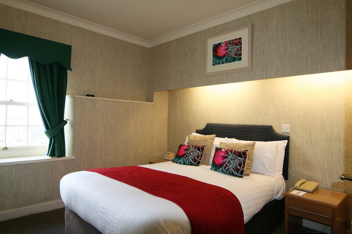 A neatly made bed with a red blanket, a bedside table with a lamp, and a window with curtains.