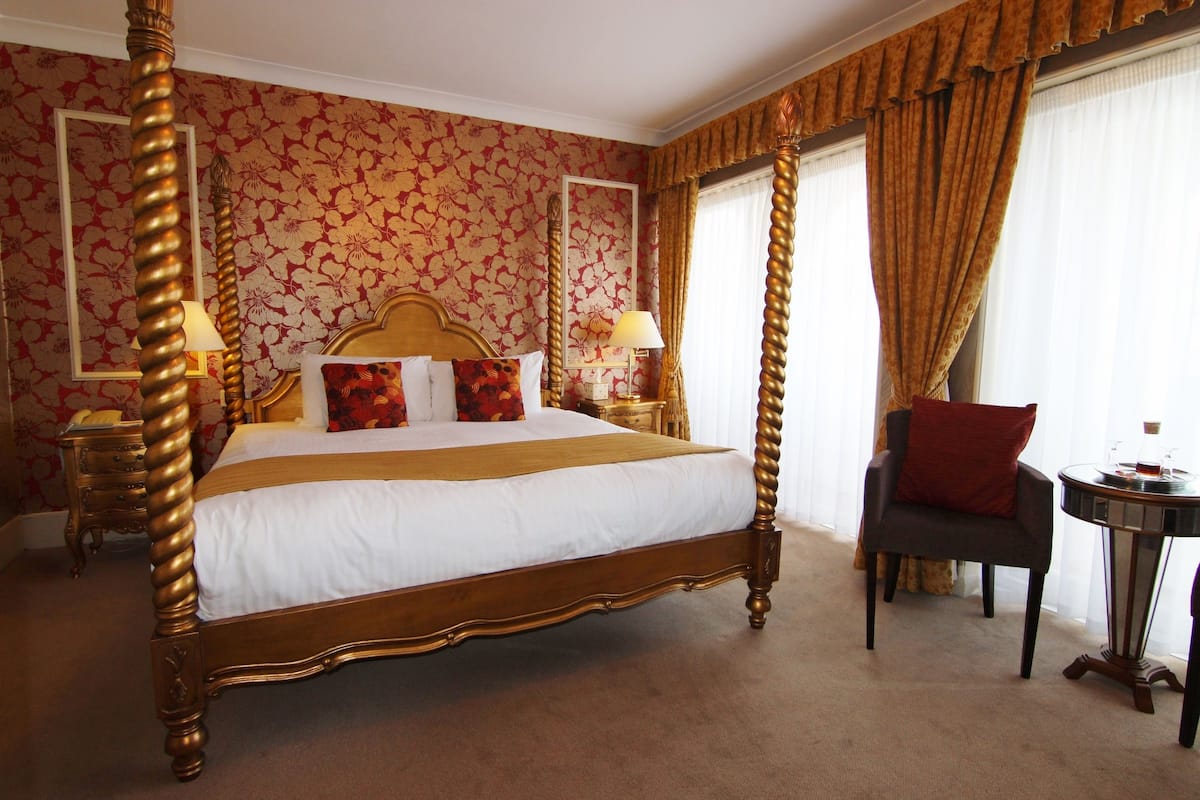A four-poster bed with a wooden frame and white bedding, a chair, a small round table, and a mirror on the wall.
