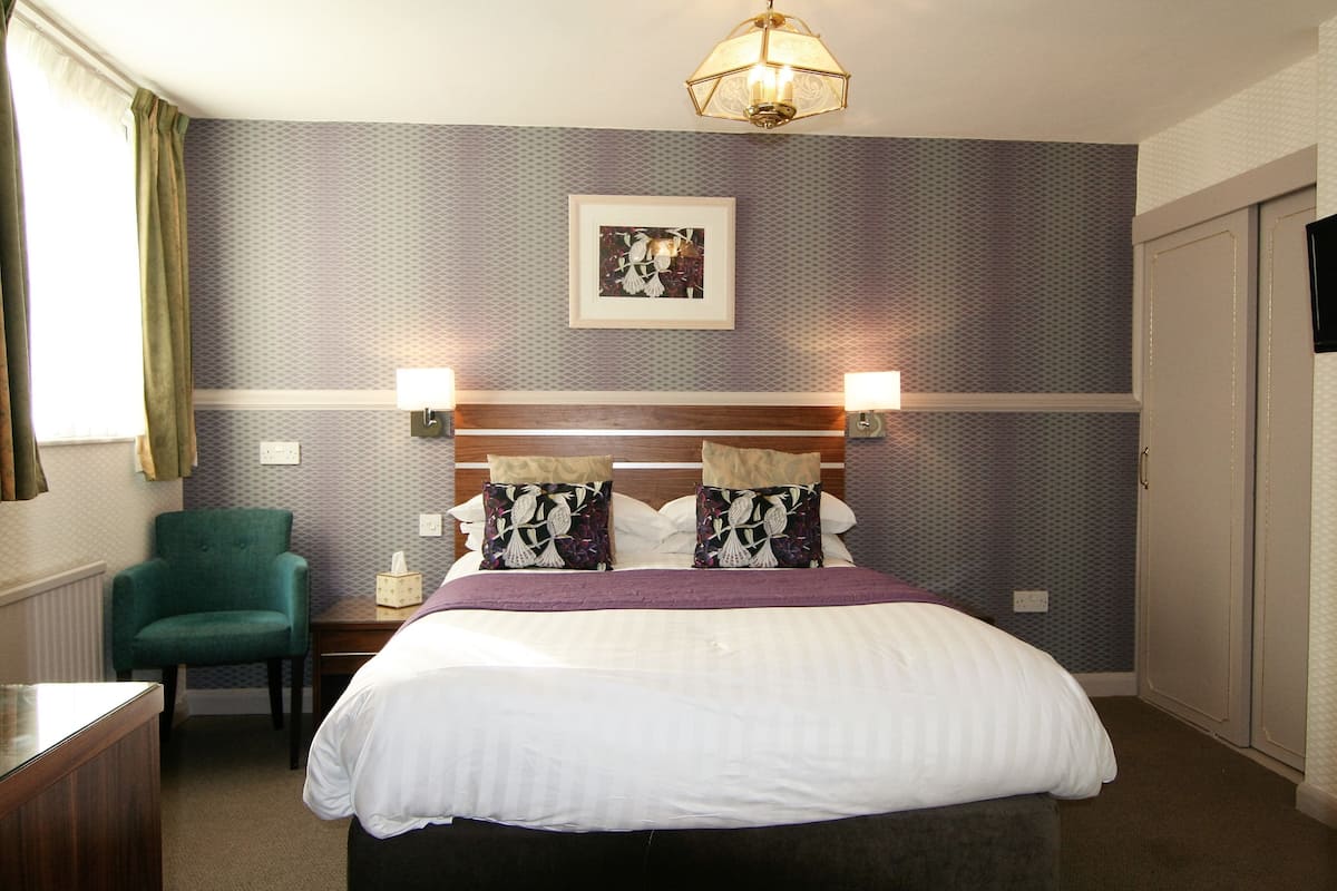 A neatly arranged hotel room with a bed, two bedside lamps, a green armchair, a bedside table, and a framed picture on the wall.