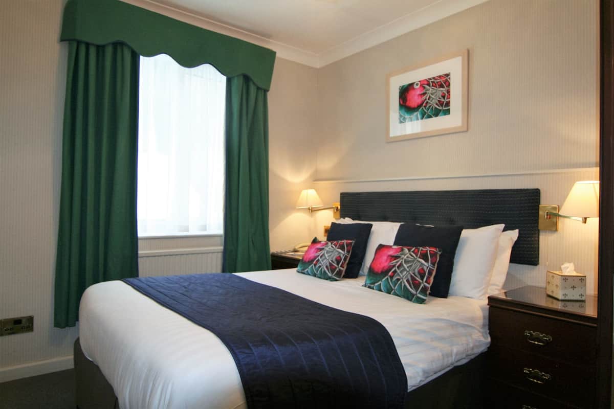 A hotel room with a large bed, green curtains, two bedside lamps, a framed artwork on the wall, and a bedside table with a tissue box.