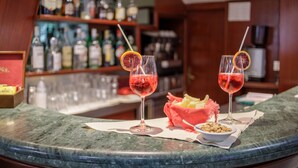 A bar counter with two glasses of red drink garnished with orange slices, a bowl of nuts, and a basket of fruit. - Hotel Astor Victoria (Forte dei Marmi)