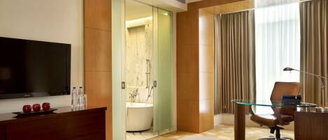 A hotel room with a large bed, a TV, a desk with a chair, a glass partition, a bathroom with a bathtub, and a large window with curtains.