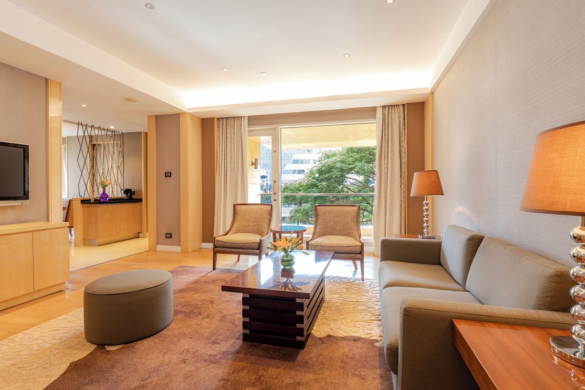 A modern hotel room with a sofa, two armchairs, a coffee table, a TV, and a balcony with a view of the city.