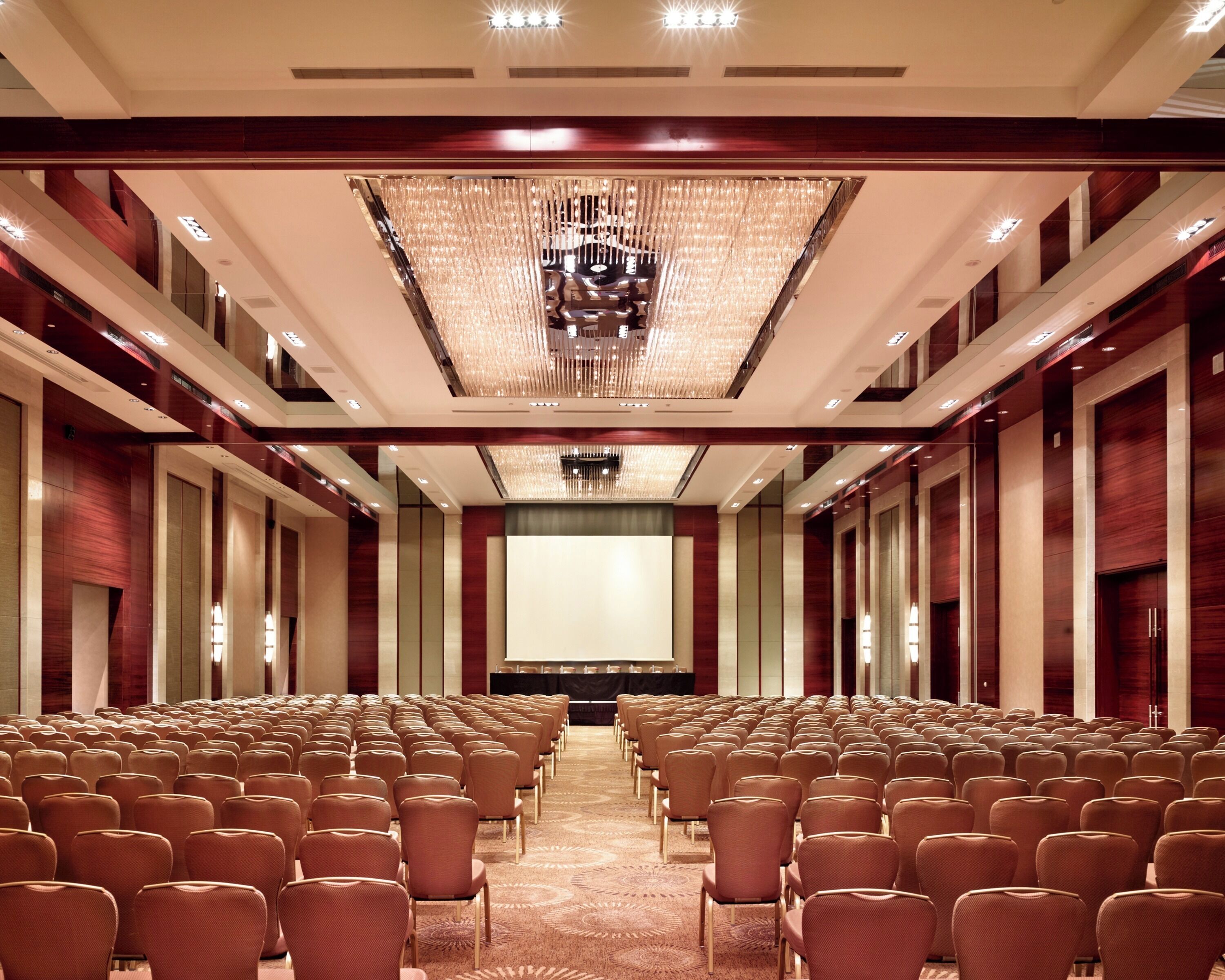 Une salle de conférence avec des rangées de chaises, un grand écran et un lustre.