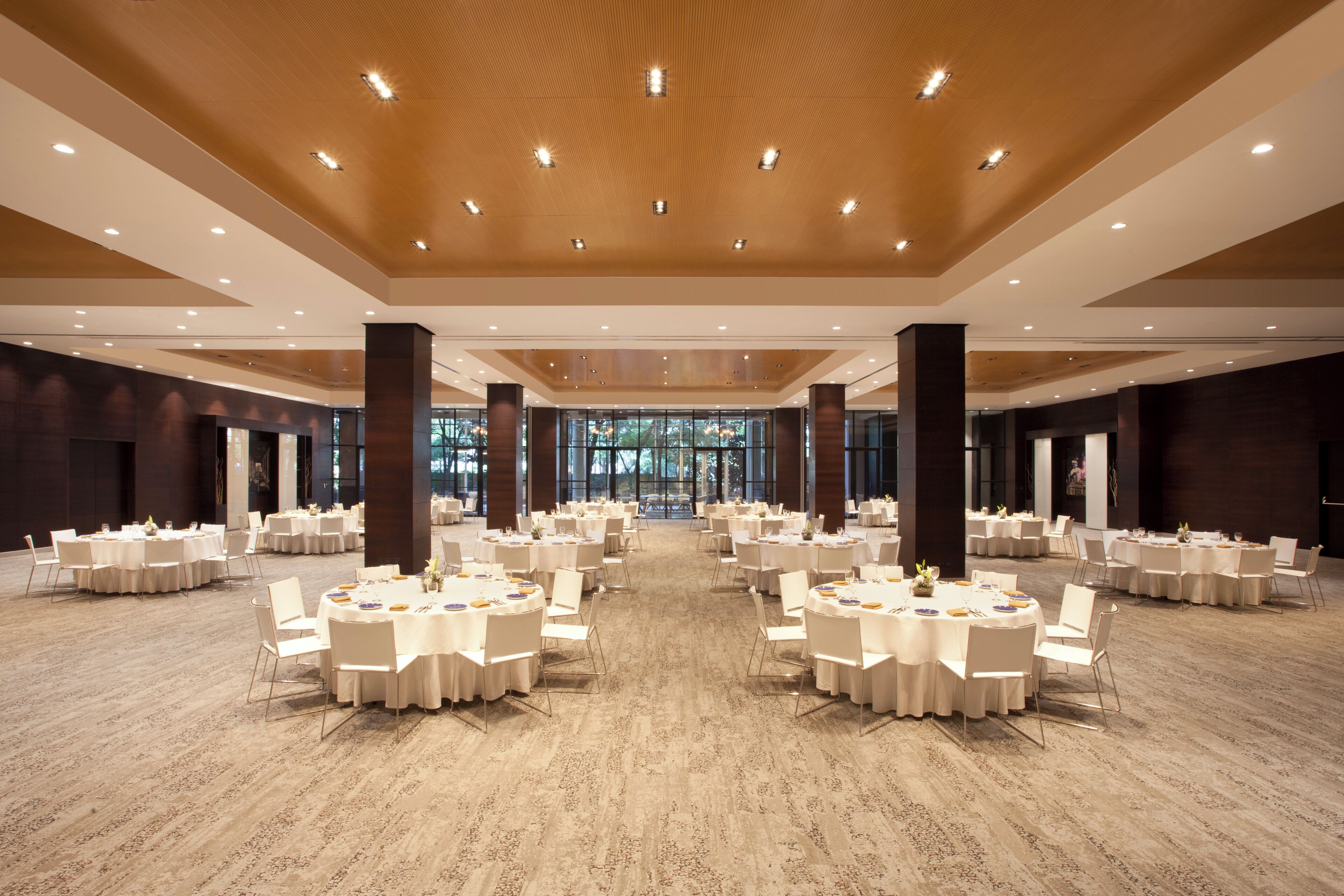 Une salle de banquet spacieuse, dotée de tables et de chaises rondes, d’un plafond en bois et de grandes fenêtres.