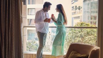 A couple standing by a window in a hotel room, with a view of buildings and a balcony outside.