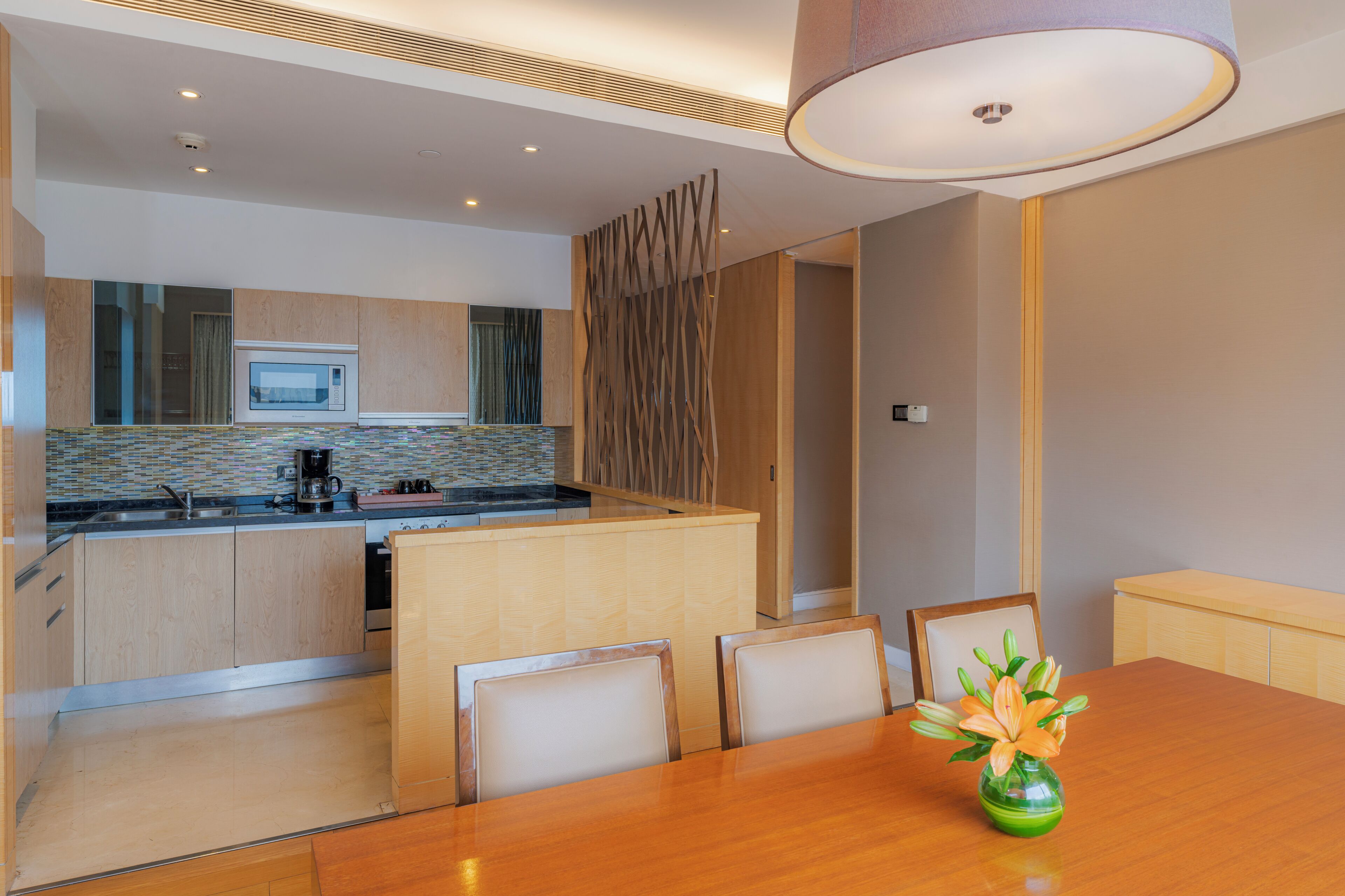 A modern kitchen with a microwave, coffee maker, and wooden cabinets. There is a dining area with a table and chairs, and a vase with flowers on the table.