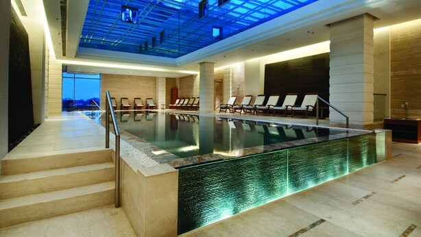 A modern indoor swimming pool with a glass ceiling, a sun terrace, and a view of the sea.