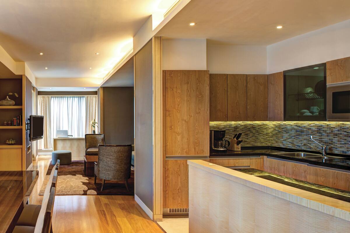 A modern hotel room with a wooden dining table, a sofa, a TV, and a kitchen area with a sink and stove.