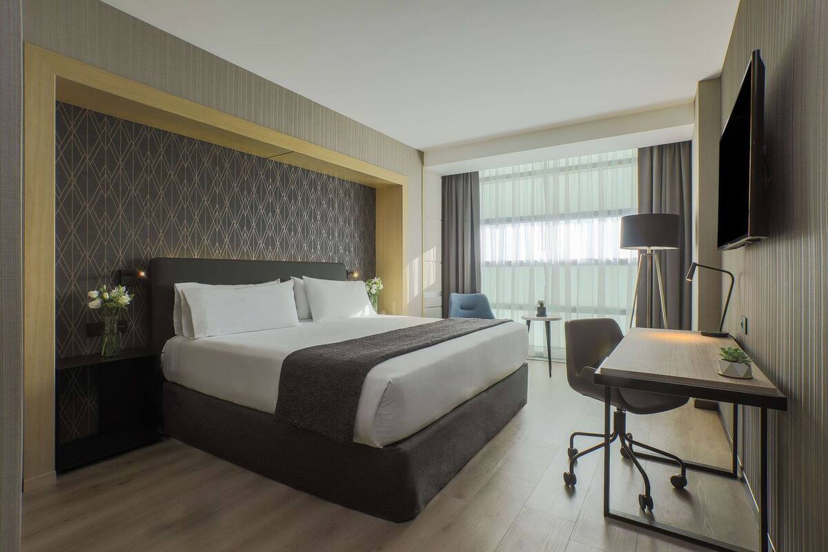 Ein Hotelzimmer mit einem großen Bett, einem Schreibtisch, einem Sessel, einem Fernseher und einem Fenster mit Vorhängen.