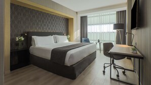 Habitación de hotel con una cama grande, un escritorio con silla, un televisor de pantalla plana y una ventana con cortinas.