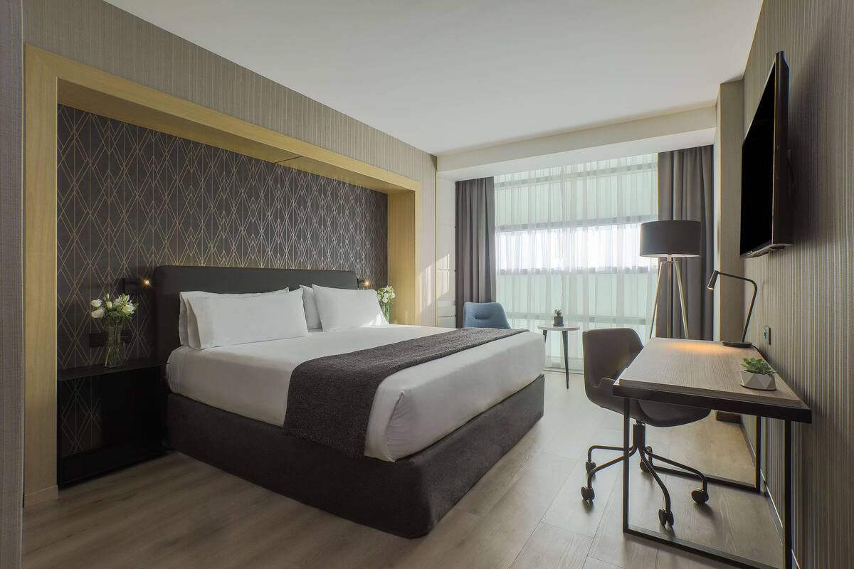 Ein Hotelzimmer mit einem großen Bett, einem Schreibtisch mit Stuhl, einem Flachbildfernseher und einem Fenster mit Vorhängen.