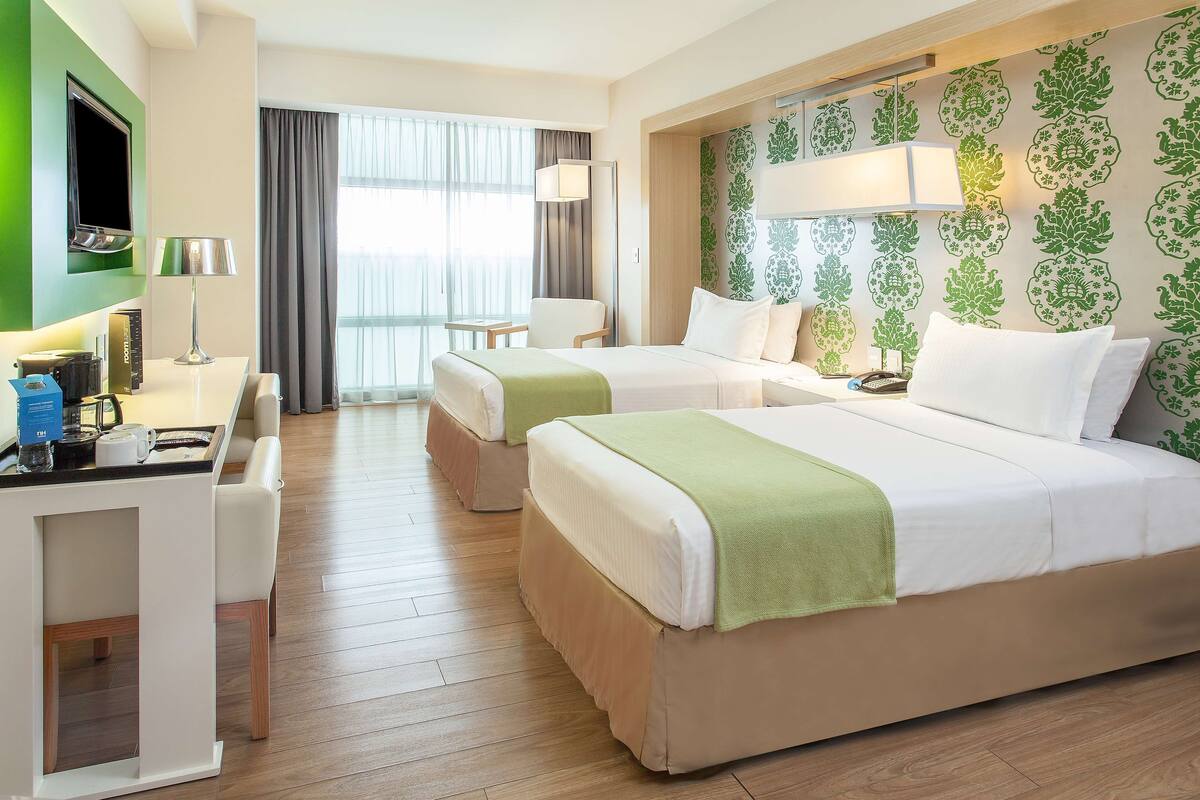 Ein Hotelzimmer mit zwei Betten, einem Schreibtisch, einem Sessel, einem Fernseher und einem großen Fenster mit Vorhängen.