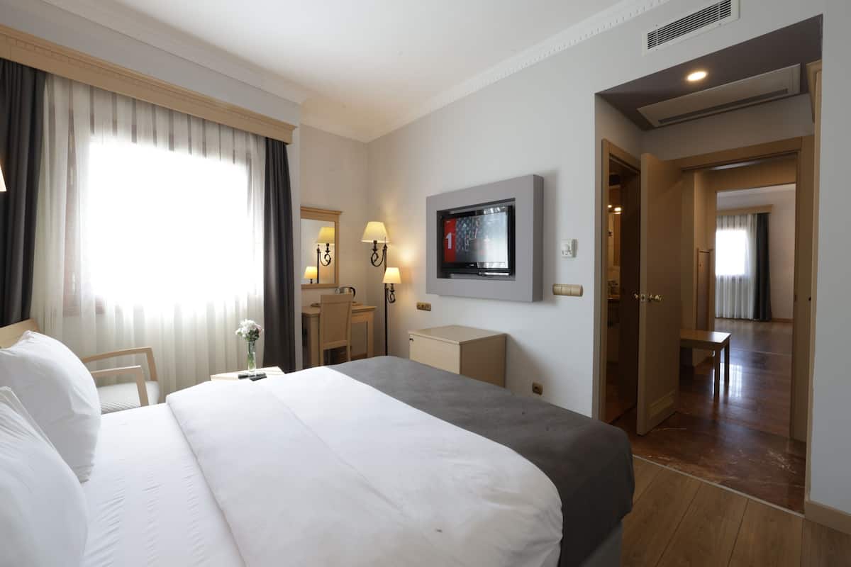 A hotel room with a large bed, a desk, a chair, a TV, and a door leading to another room.