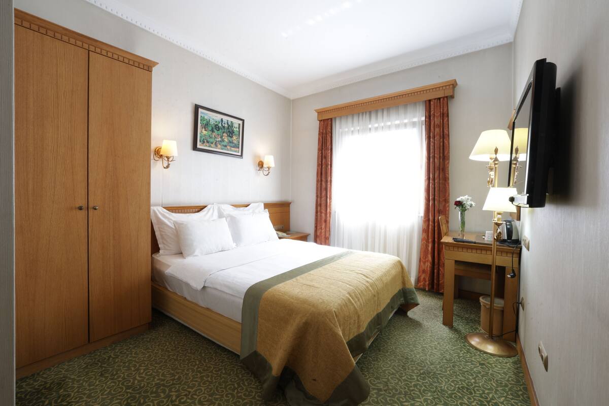 A hotel room with a bed, a wooden wardrobe, a desk with a lamp, and a television.