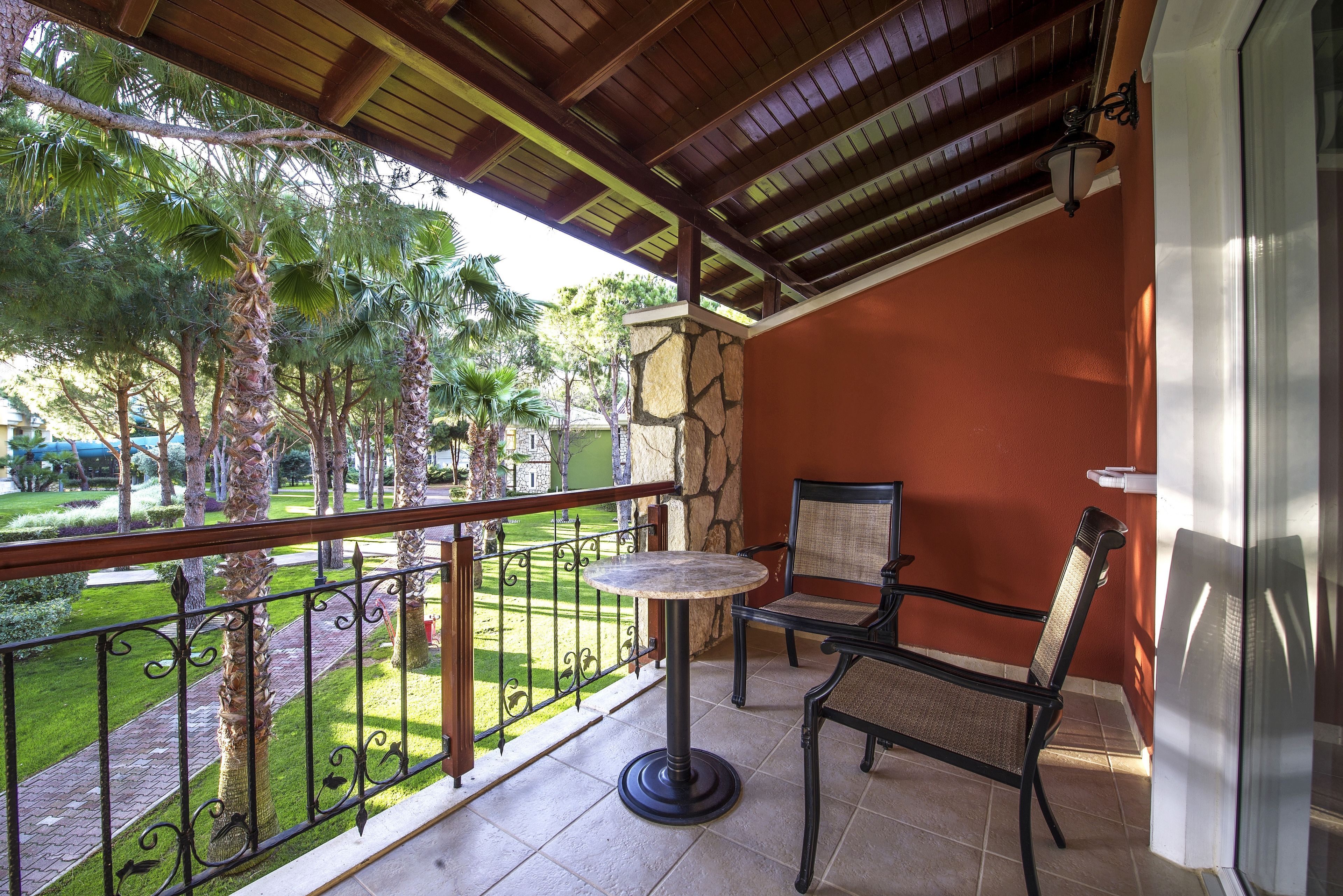 A balcony with a table and chair, a stone wall, and a view of a well-maintained garden with palm trees.