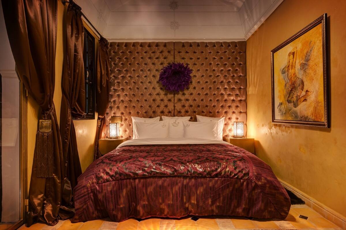 Un dormitorio con cabecera capitonada, una cama grande con colcha estampada, dos lámparas de noche y un cuadro en la pared.