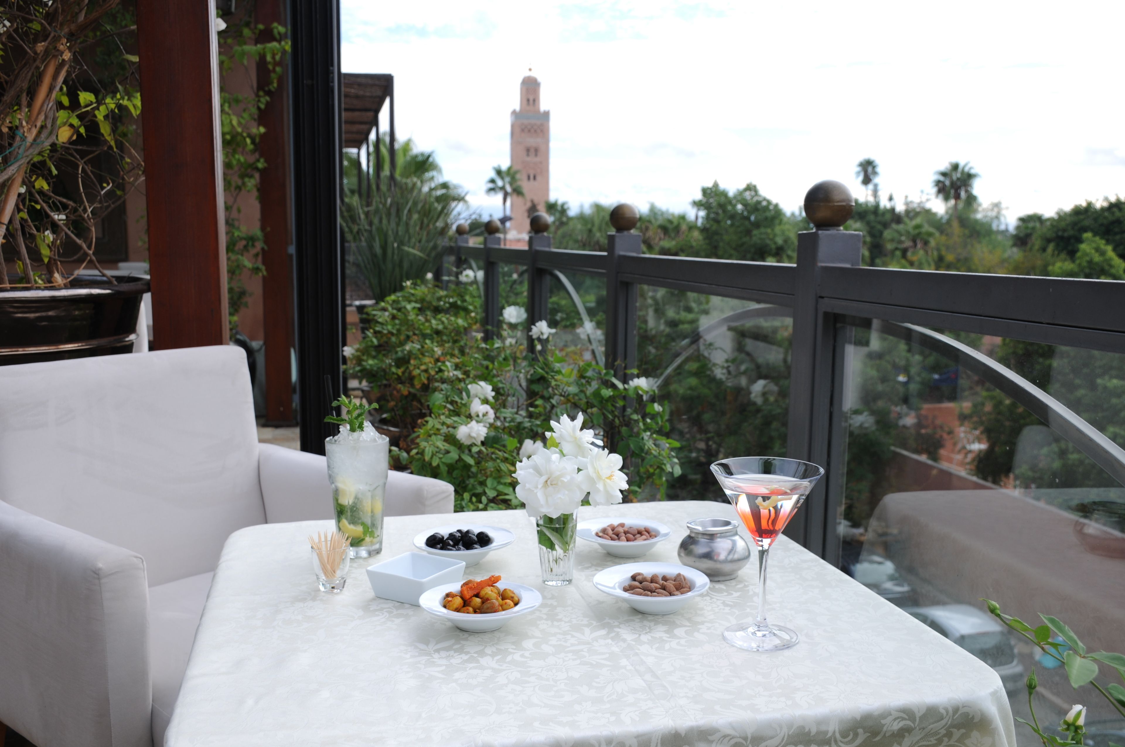 Mesa posta com diversos pratos, uma taça de martini e um vaso de flores brancas, com vista para um jardim e uma torre ao longe.