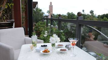 Mesa posta com diversos pratos, uma taça de martini e um vaso de flores brancas, com vista para um jardim e uma torre ao longe.