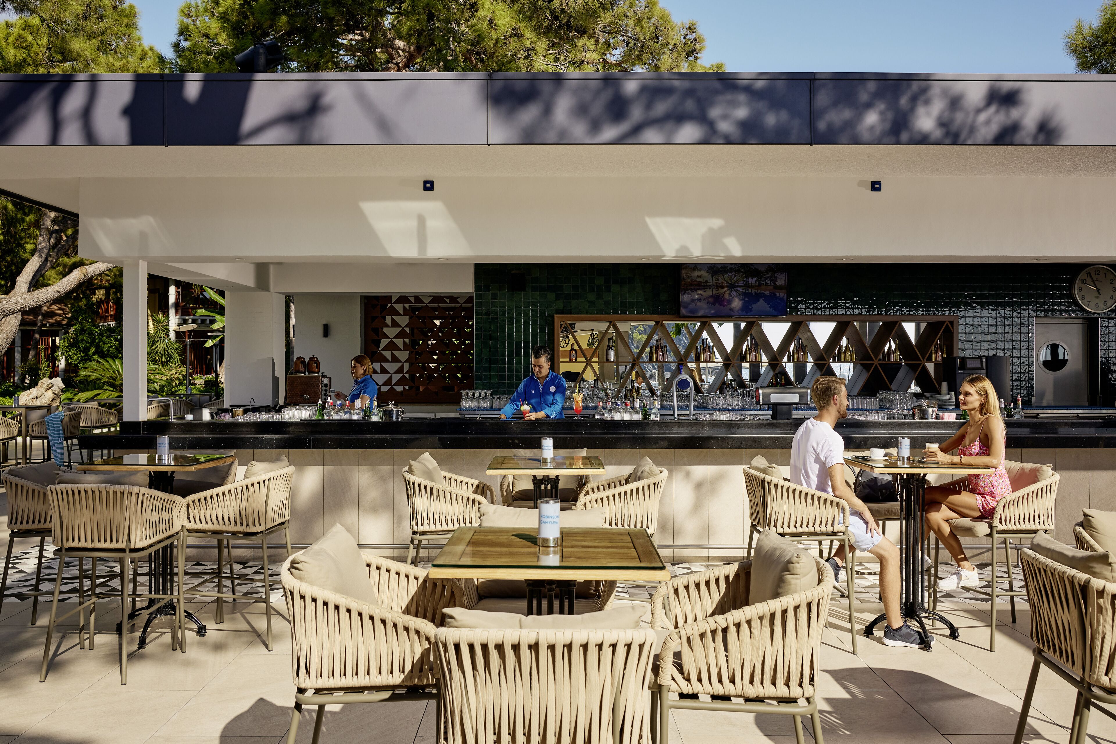 Un espace bar extérieur avec des chaises et des tables en osier, un barman et une horloge au mur.