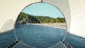 A view from a boat's deck with a netted railing, a clear blue sea, and a beach with trees and umbrellas in the background. - ROBINSON ÇAMYUVA Adults Only- All Inclusive (Kemer)