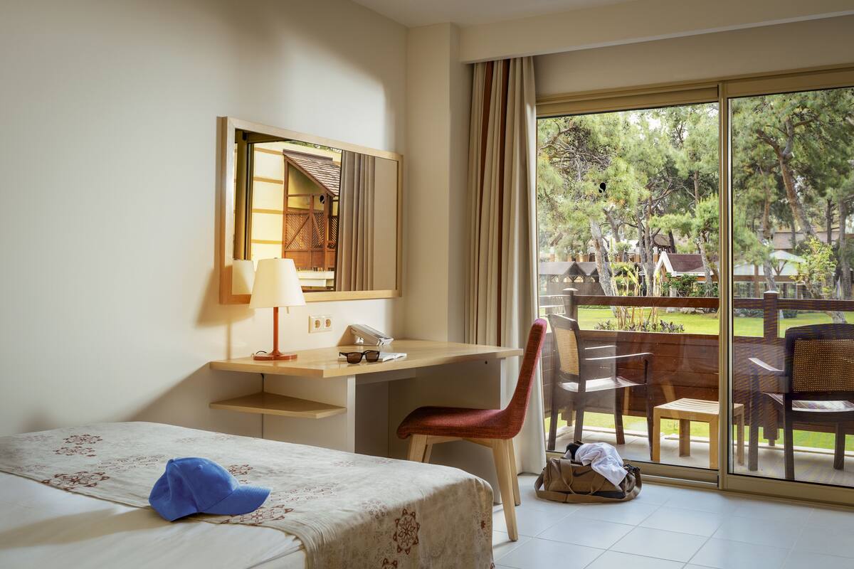 A hotel room with a bed, a desk, a chair, a lamp, and a view of the outdoors.