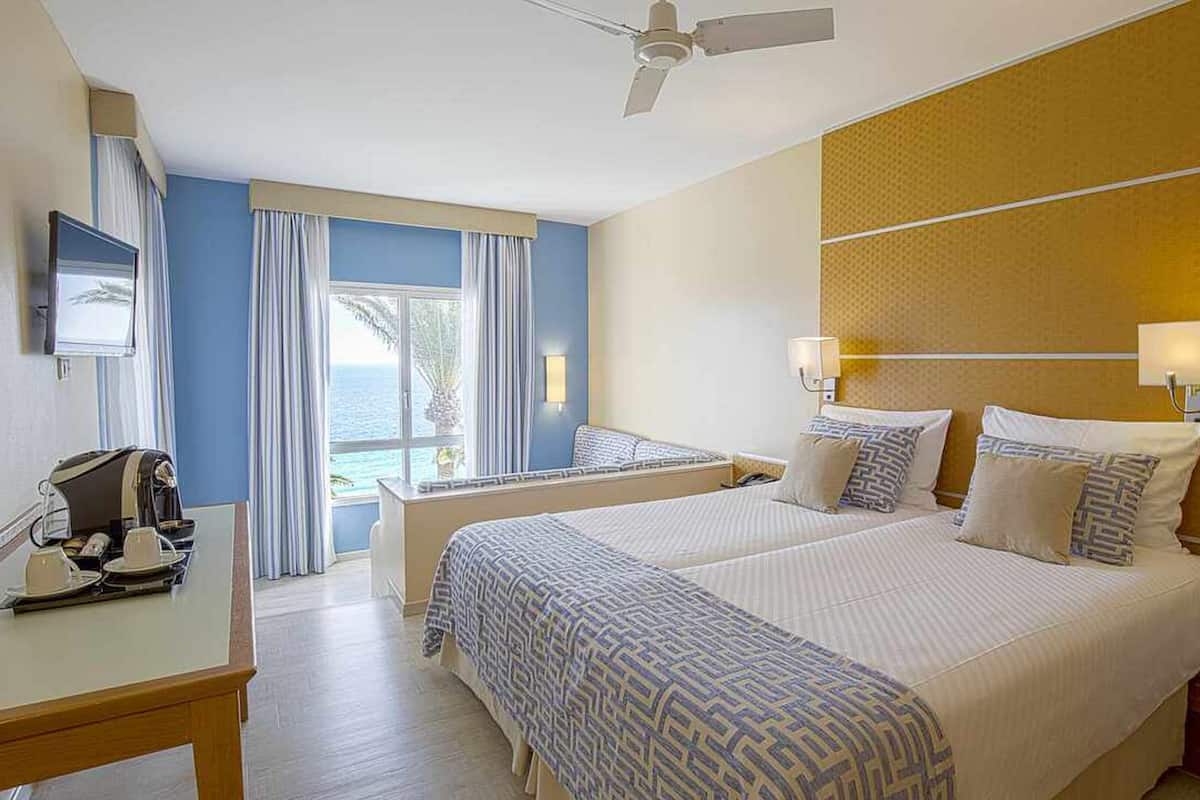 Une chambre d’hôtel avec deux lits, un bureau avec une machine à café, une télévision et un balcon avec vue sur la mer.
