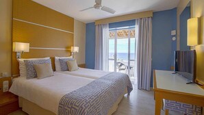 A hotel room with two beds, a desk, a chair, a ceiling fan, and a balcony with a view of the sea. - ROBINSON ESQUINZO PLAYA - All Inclusive (Pajara)