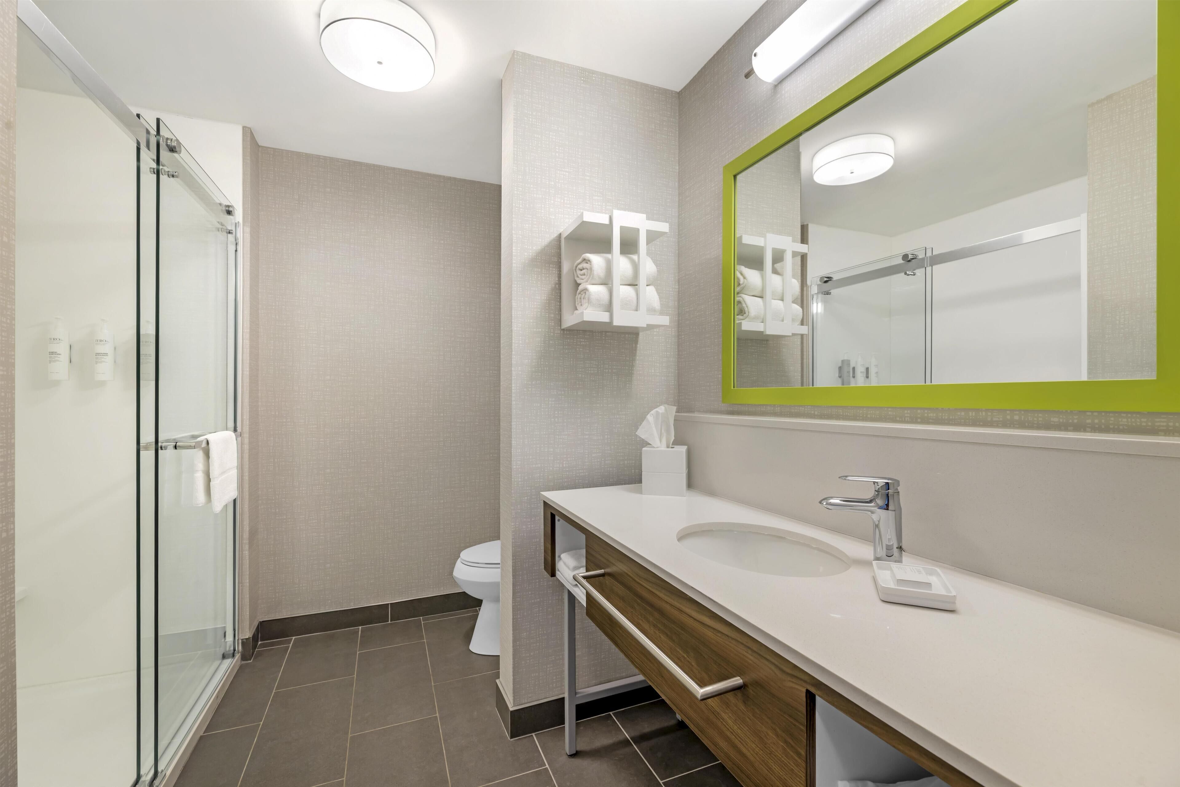 A modern bathroom with a glass shower enclosure, a white sink with a wooden vanity, and a mirror with a green frame.