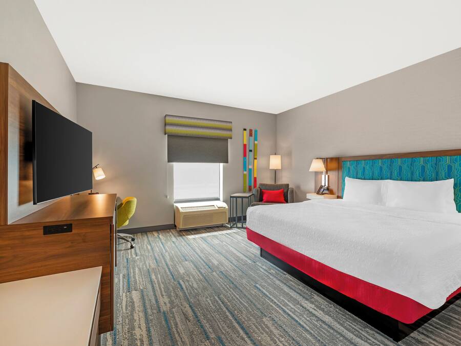A hotel room with a large bed, a flat-screen TV, a desk, a chair, a window with blinds, and a colorful wall decoration.