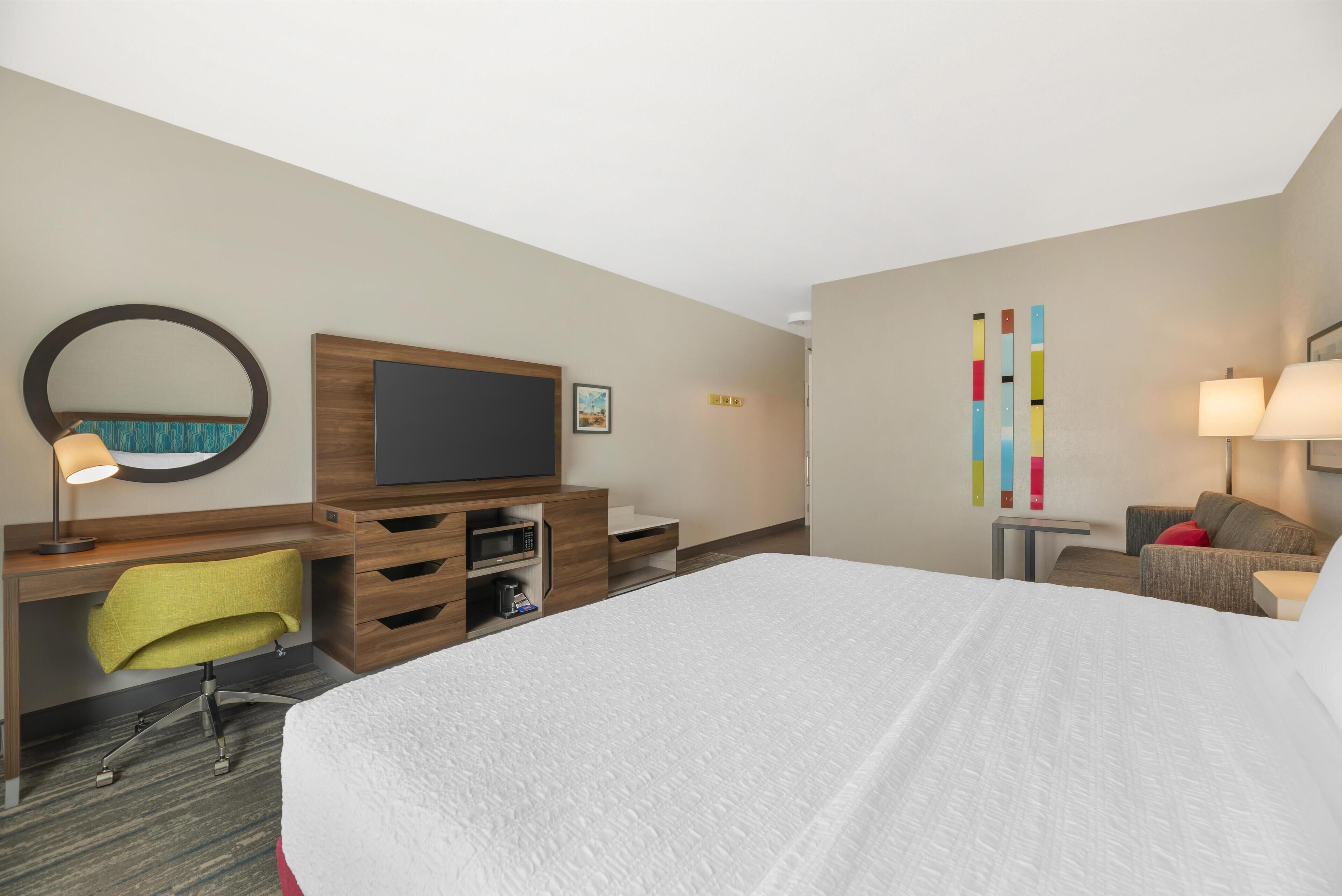Une chambre d’hôtel avec un grand lit, un bureau équipé d’un ordinateur et d’un téléviseur à écran plat, une chaise, une petite table avec une lampe, et une œuvre d’art abstraite et colorée accrochée au mur.