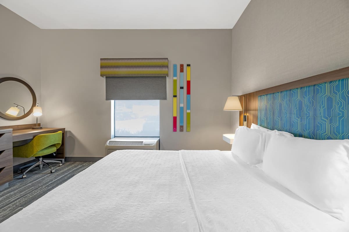 A hotel room with a large bed, a desk with a chair, a window with a view, and a wall with a geometric pattern.