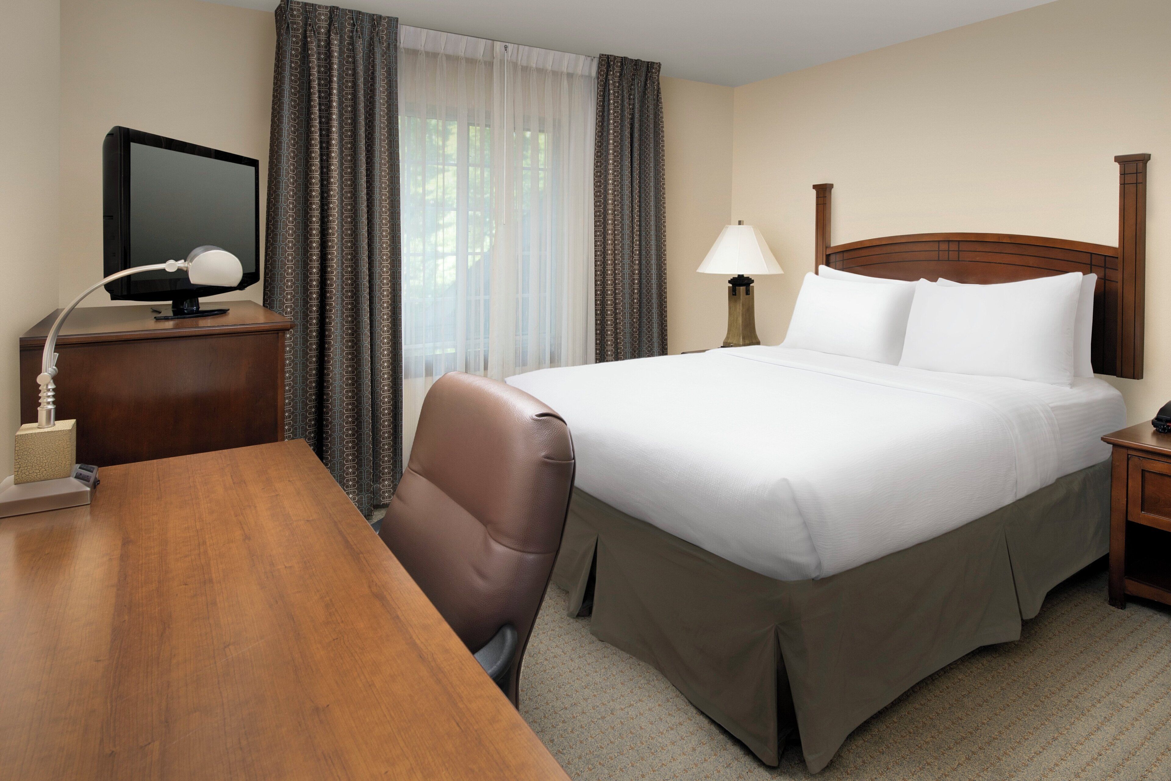 A hotel room with a bed, a desk, a chair, a TV, and a lamp.