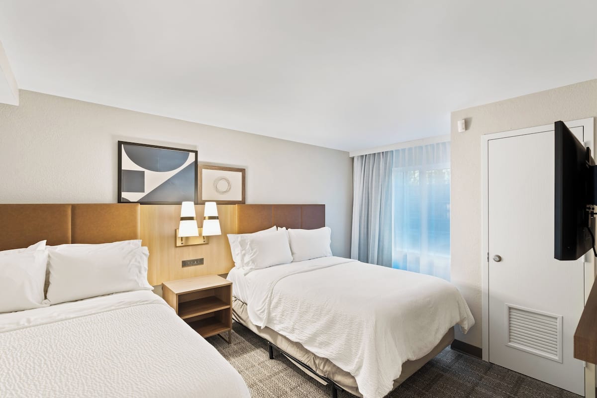 A hotel room with two beds, a nightstand with lamps, a TV mounted on the wall, and a window with curtains.