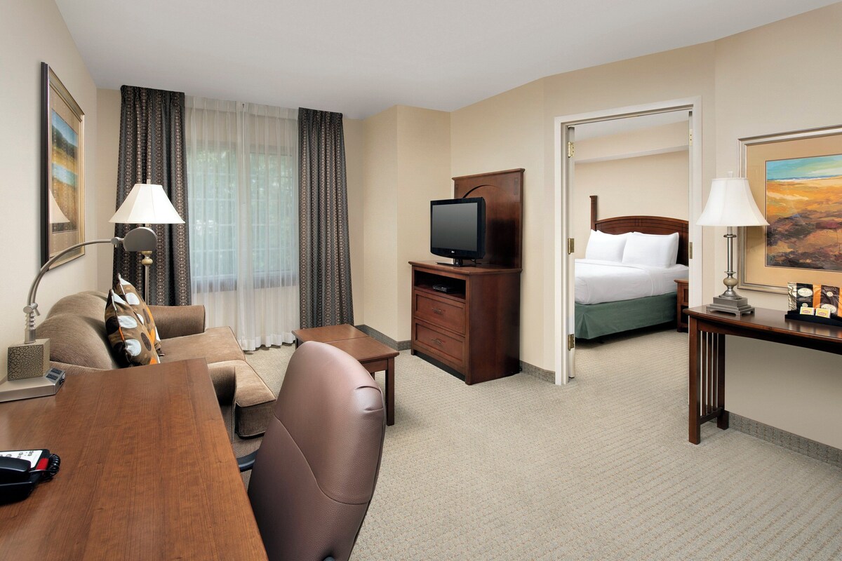 A hotel room with a bed, a desk, a television, a lamp, and a painting on the wall.
