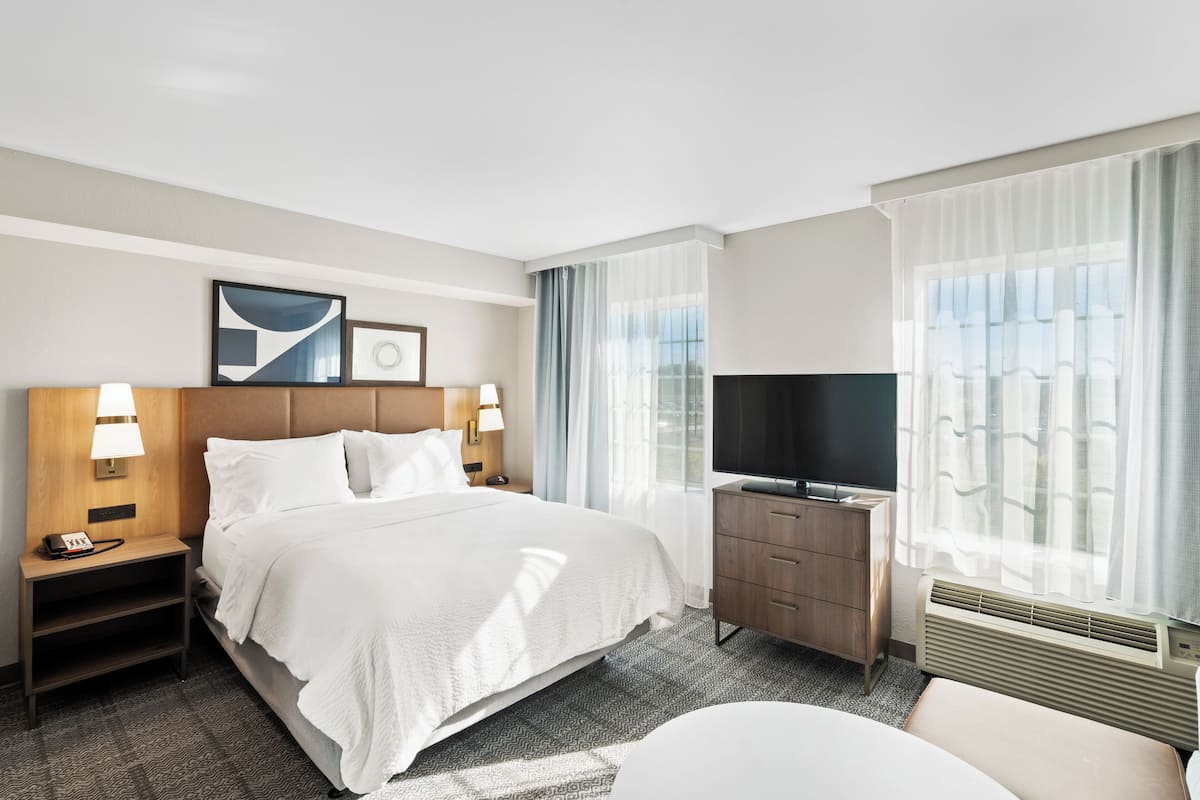 A hotel room with a large bed, two bedside tables with lamps, a dresser, a television, and a window with curtains.