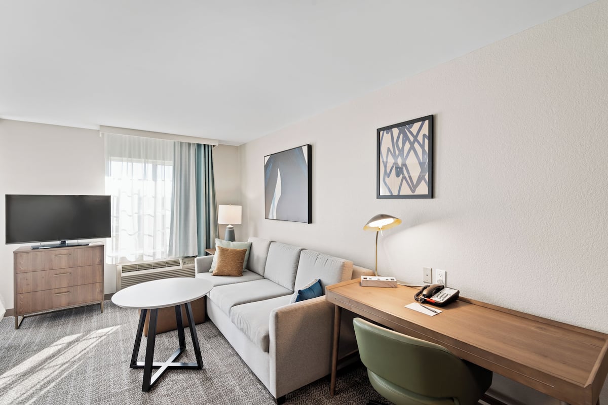 A modern hotel room with a sofa, a small round table, a desk with a chair, a television, and artwork on the walls.