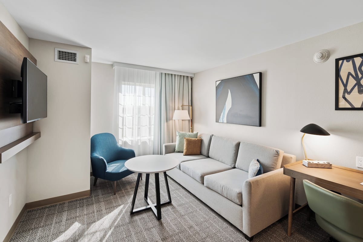 A modern hotel room with a sofa, a small round table, a desk with a lamp, a TV mounted on the wall, and a large window with curtains.