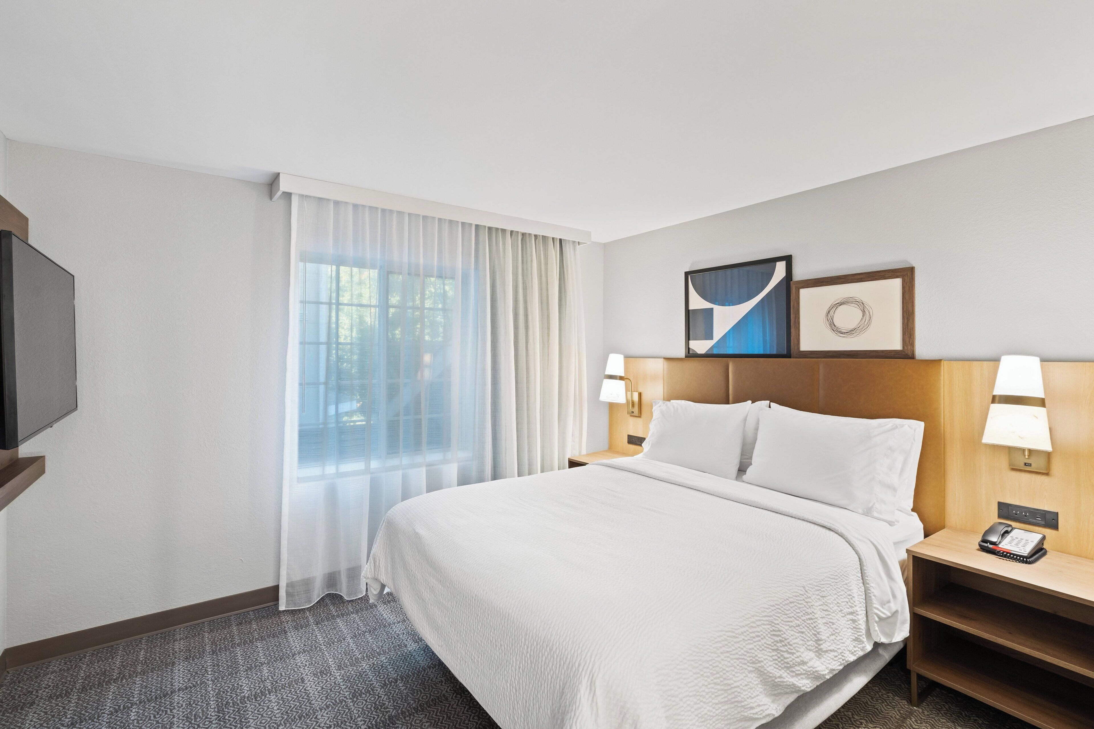 A hotel room with a large bed, a TV on the left, a window with sheer curtains, two bedside lamps, and a framed picture above the bed.