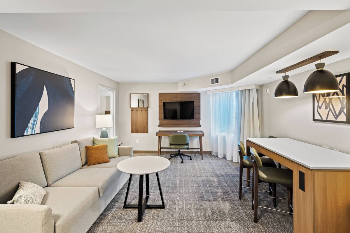 A modern hotel room with a sofa, a small round table, a desk with two chairs, a TV, and artwork on the walls.