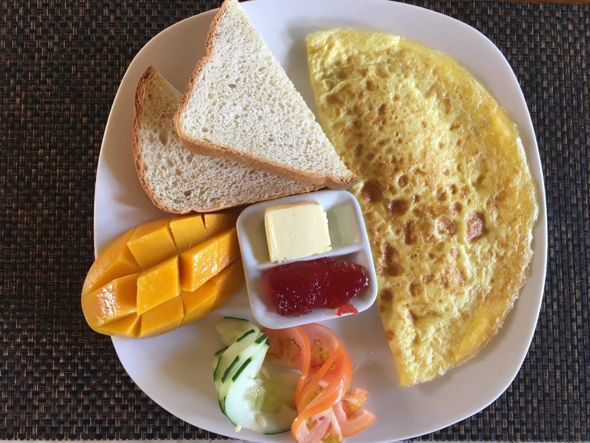 A plate with an omelette, two slices of white bread, a block of butter, mango, cucumber, carrot, and jam.