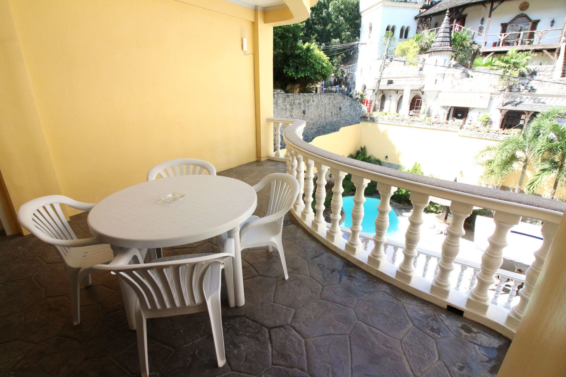 A balcony with a round table and four chairs, a stone floor, and a view of a pool and buildings in the background.