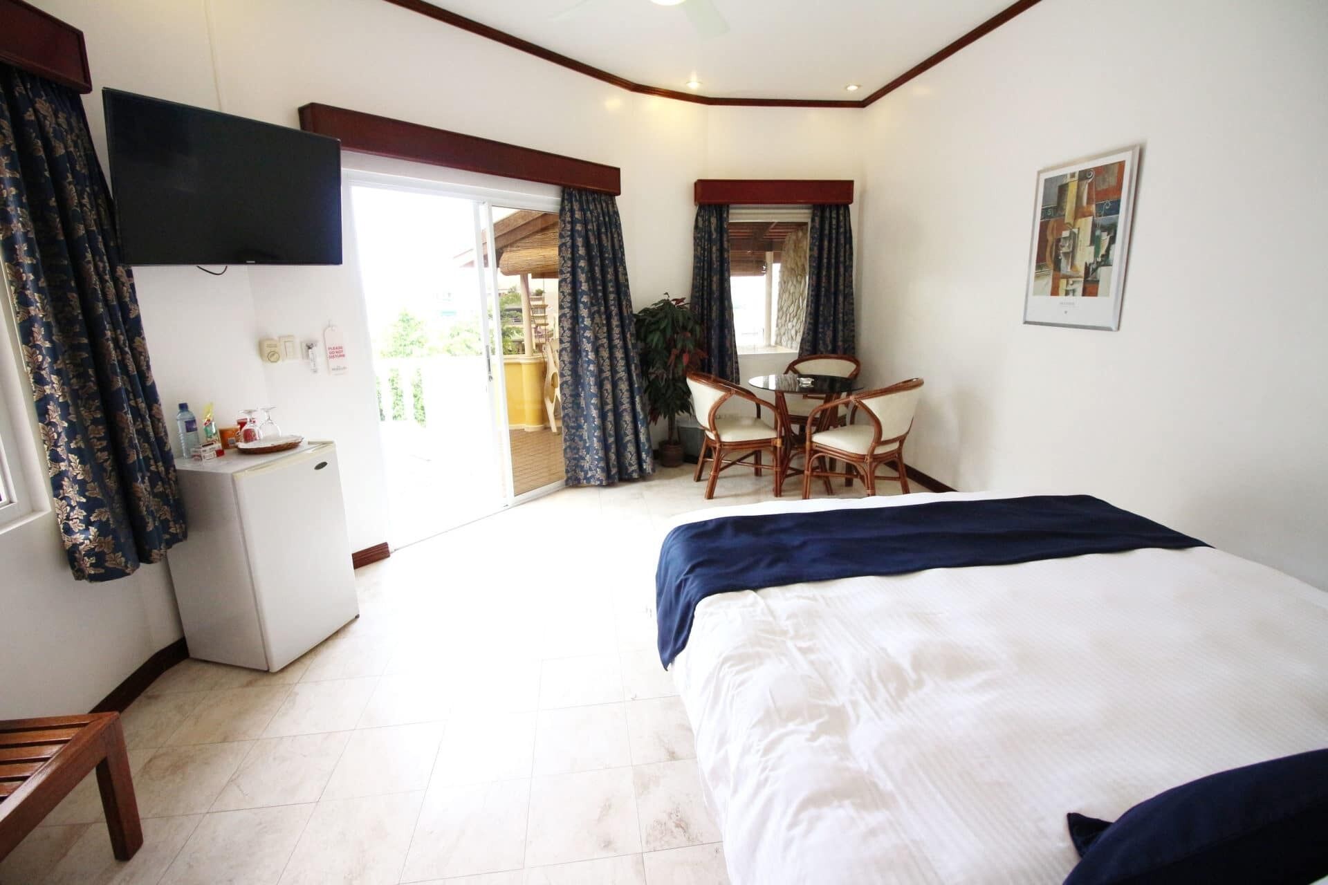 A hotel room with a bed, a desk, and a chair. There is a television mounted on the wall, and a window with curtains.