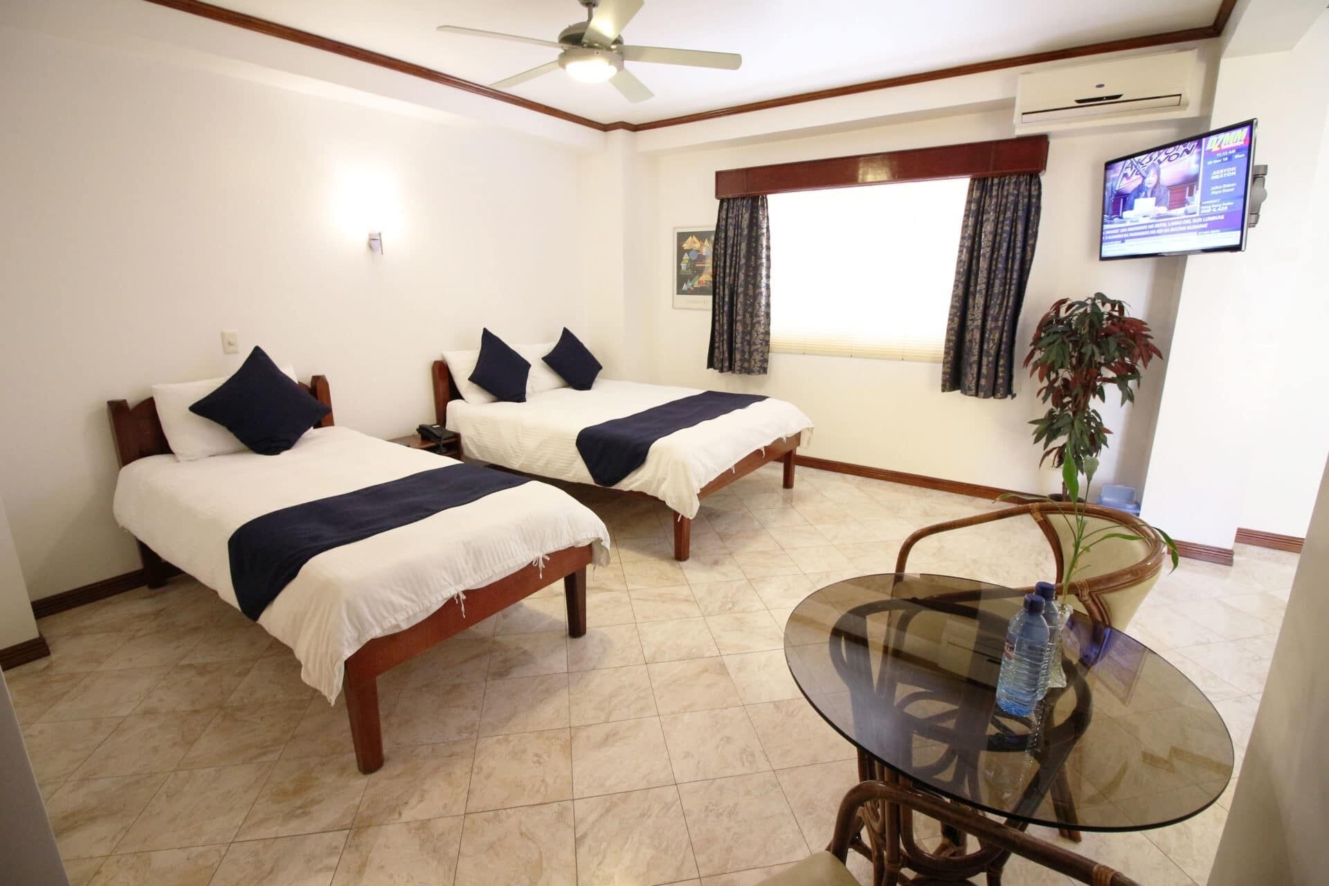 A hotel room with two beds, a glass table, a ceiling fan, a TV, and a window with curtains.