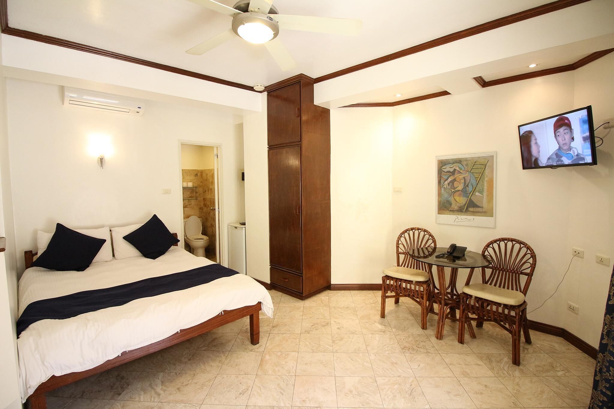 A bedroom with a bed, a ceiling fan, a TV, and a dining area with chairs.