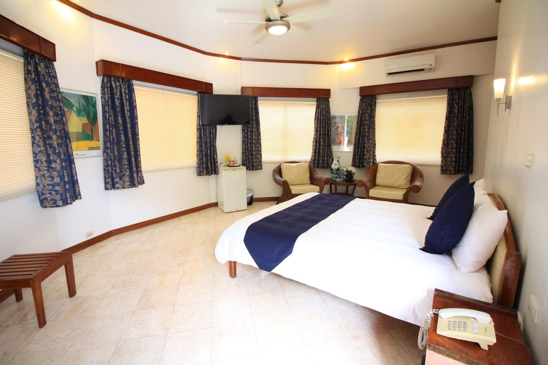 A hotel room with a large bed, a desk with a telephone, a chair, a television, and a ceiling fan.