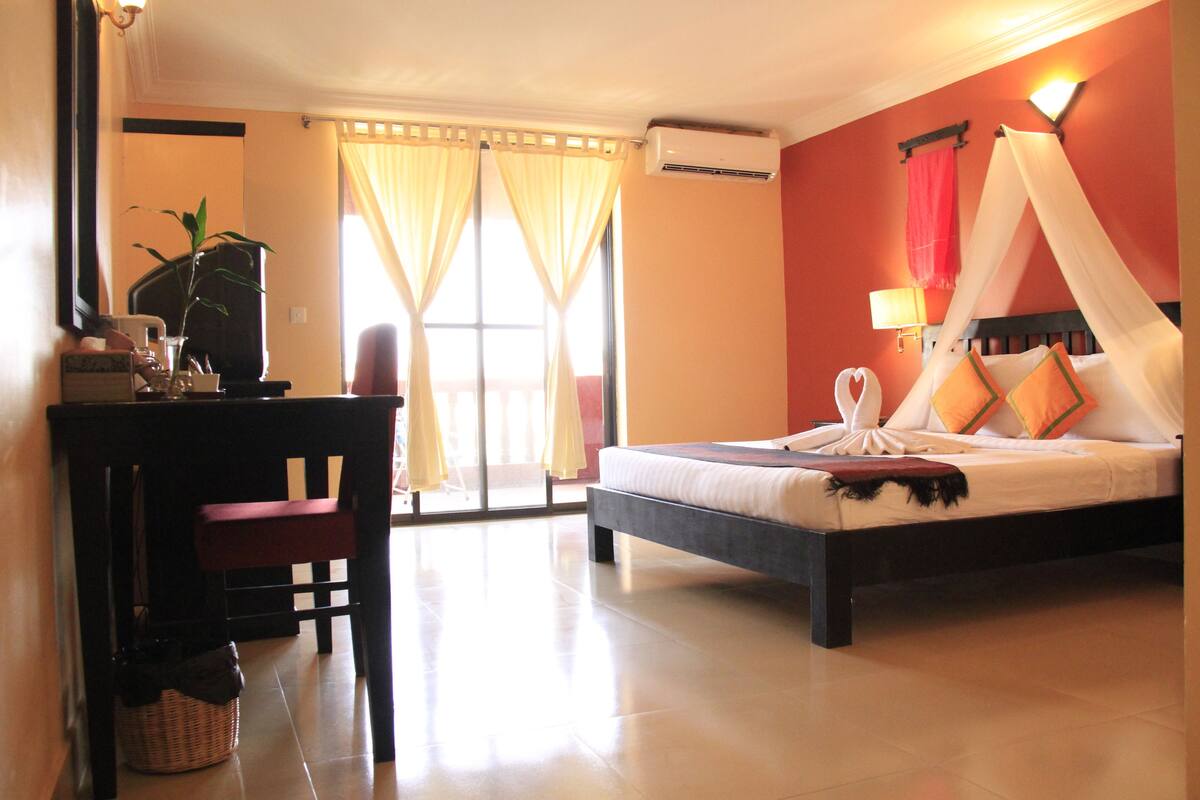 Habitación de hotel con cama, escritorio, silla, planta en maceta y vistas al exterior.
