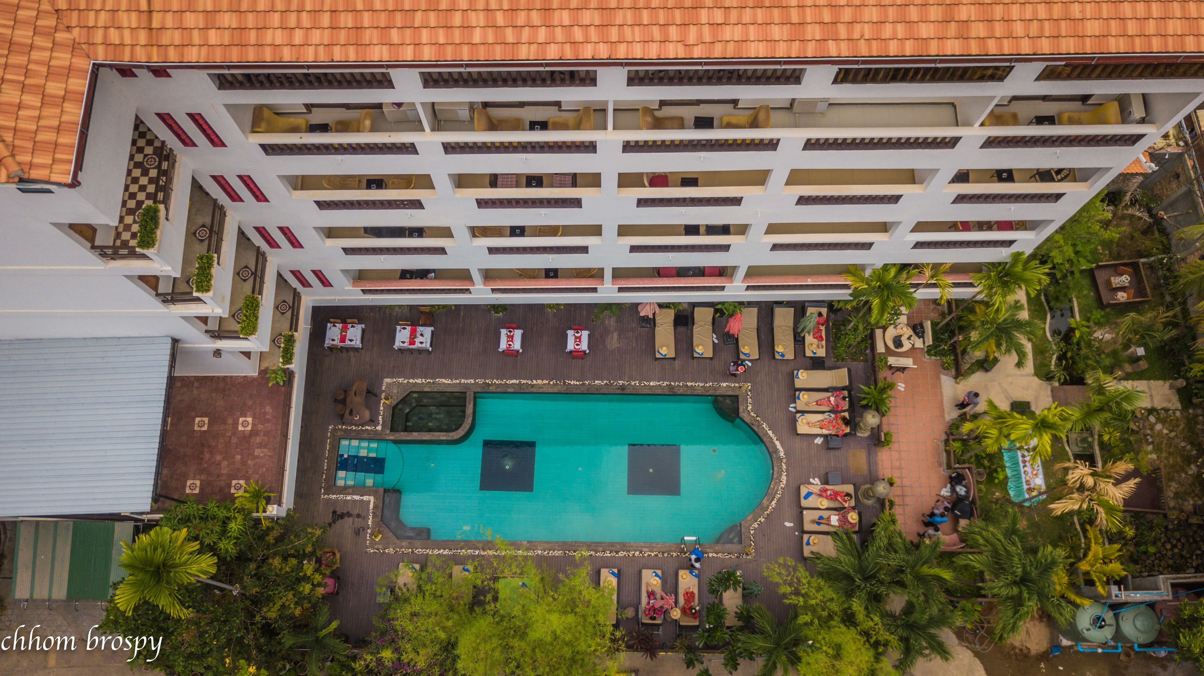 An aerial view of a hotel with a swimming pool surrounded by lounge chairs and sunbeds, a covered patio area, and a rooftop terrace.
